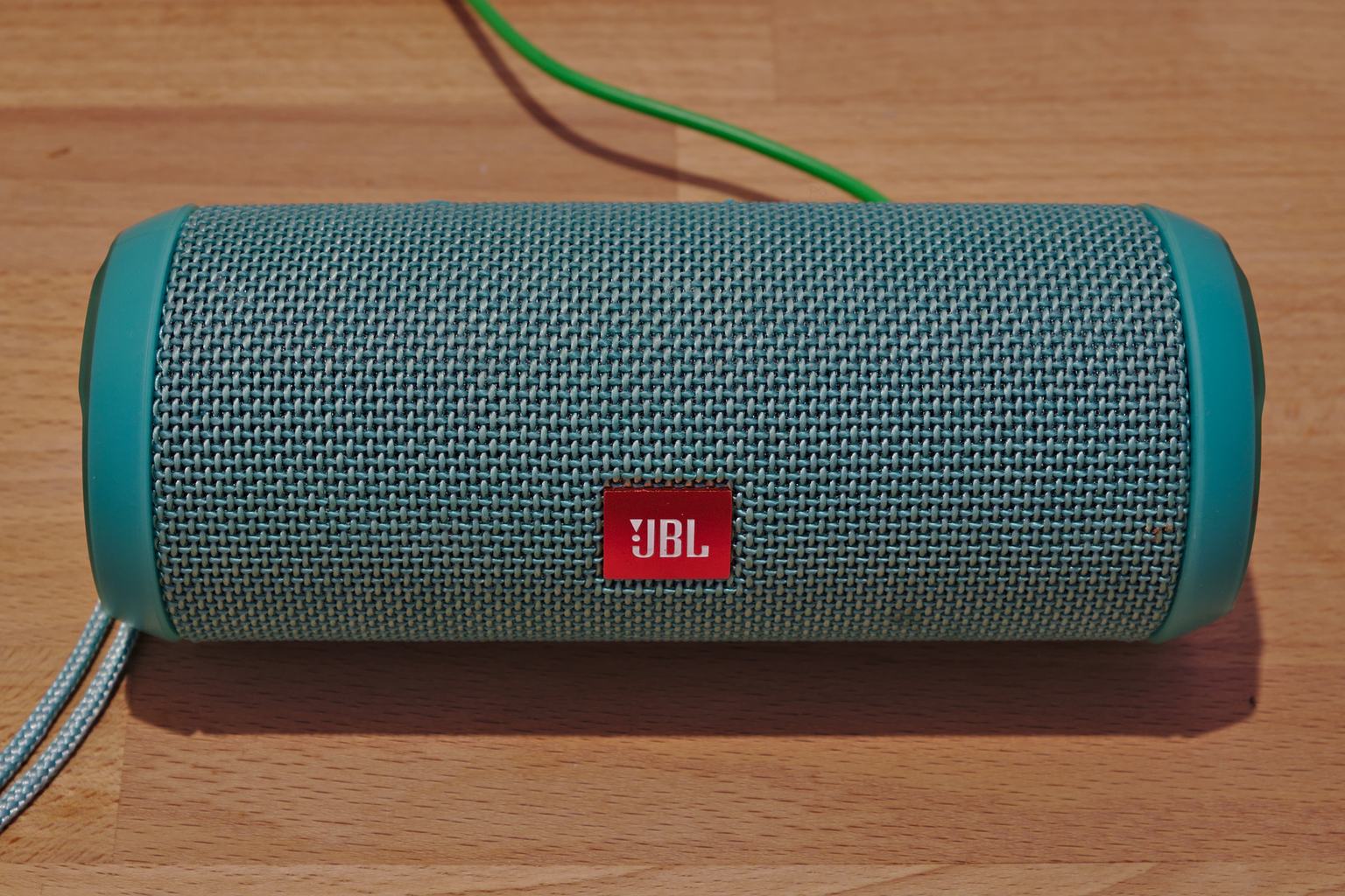 A teal JBL Flip 3 portable Bluetooth speaker lying on a wooden table, with its woven grille and red JBL badge facing the camera