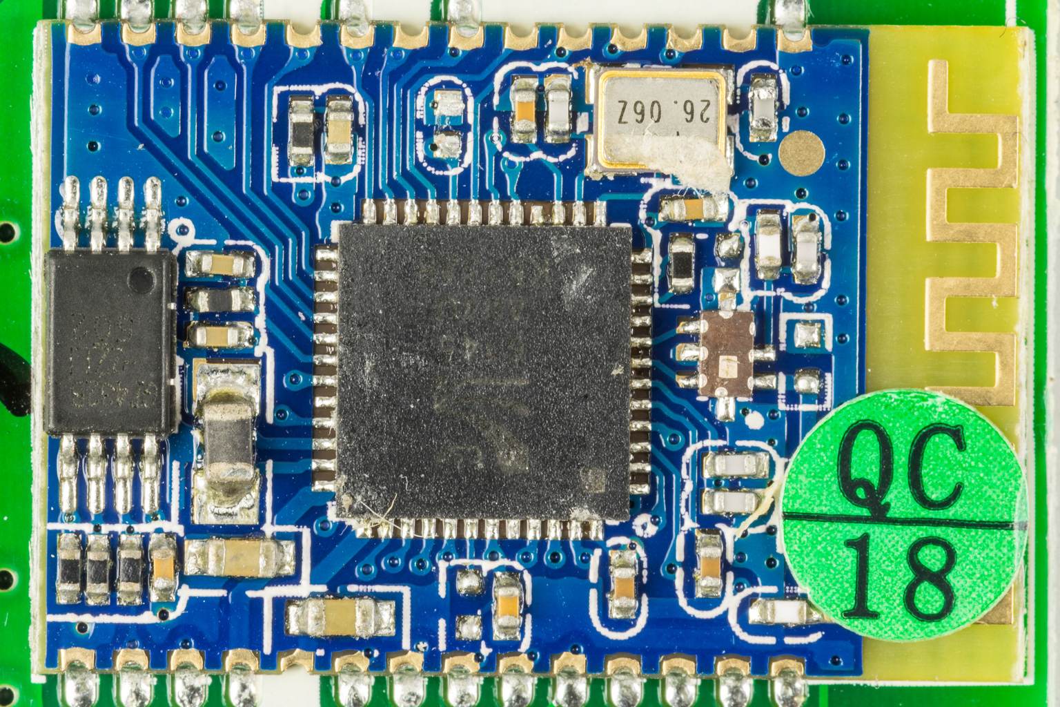 Macro photograph of a Bluetooth radio module soldered to a circuit board, showing the shielded system-on-chip, a 26.062 MHz crystal, the chip antenna on the right edge and a green quality-control sticker