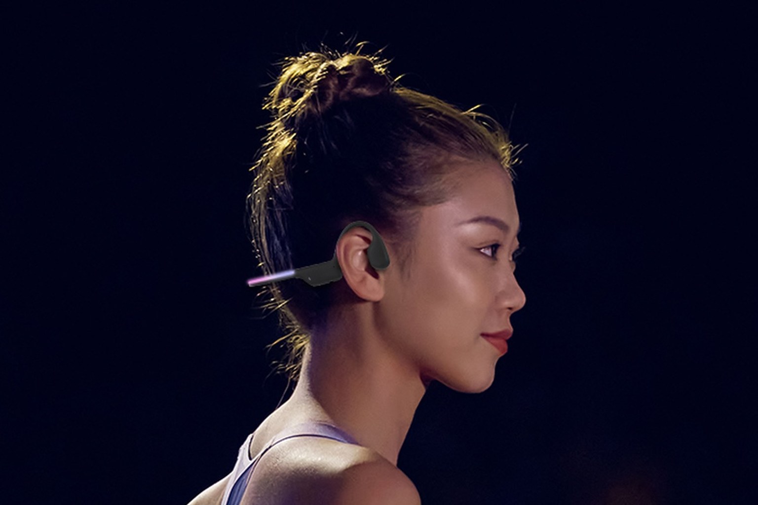 A runner at night wearing a bone conduction headband with the transducer on the cheekbone, ears fully open, city lights behind