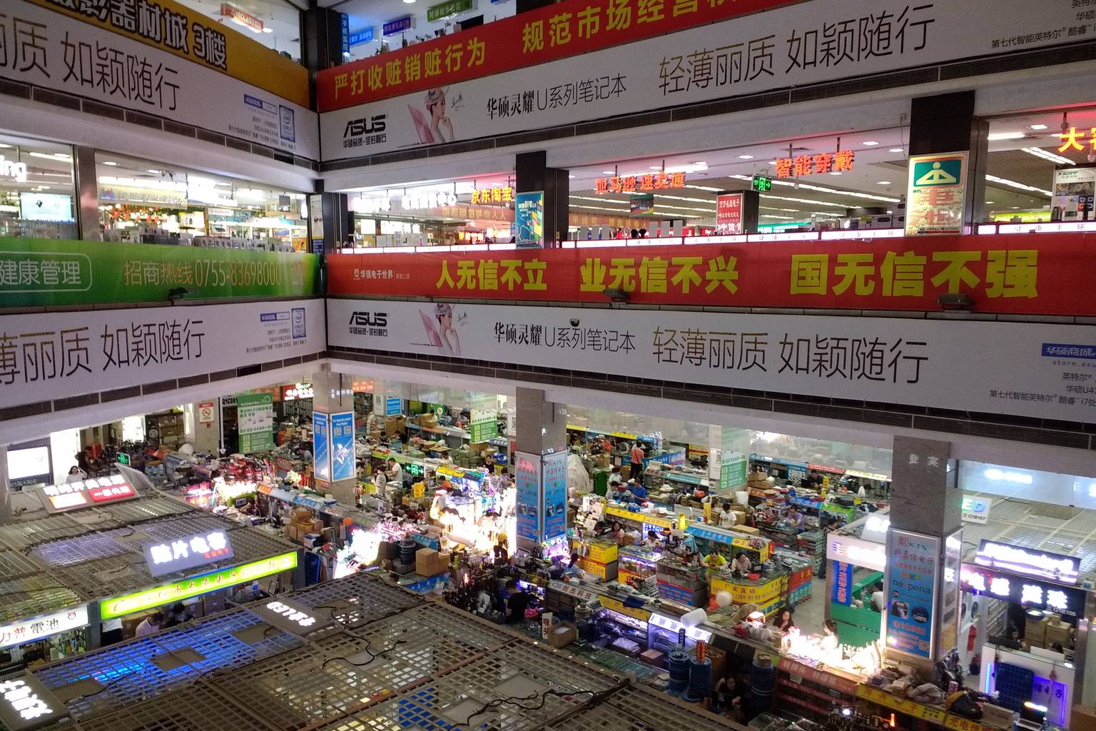 Interior of the Huaqiangbei electronics market in Shenzhen, seen from a balcony: two floors of small counters, stalls and stacked stock under large advertising banners