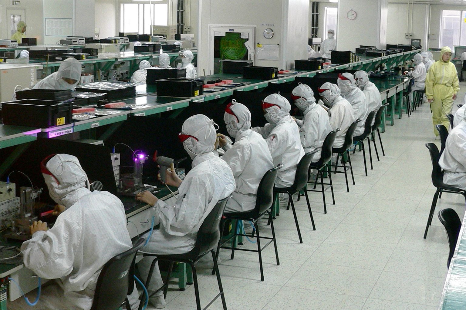 A long electronics assembly hall in Shenzhen, workers in white protective suits seated along benches on both sides of a central aisle while a supervisor in a yellow suit walks past