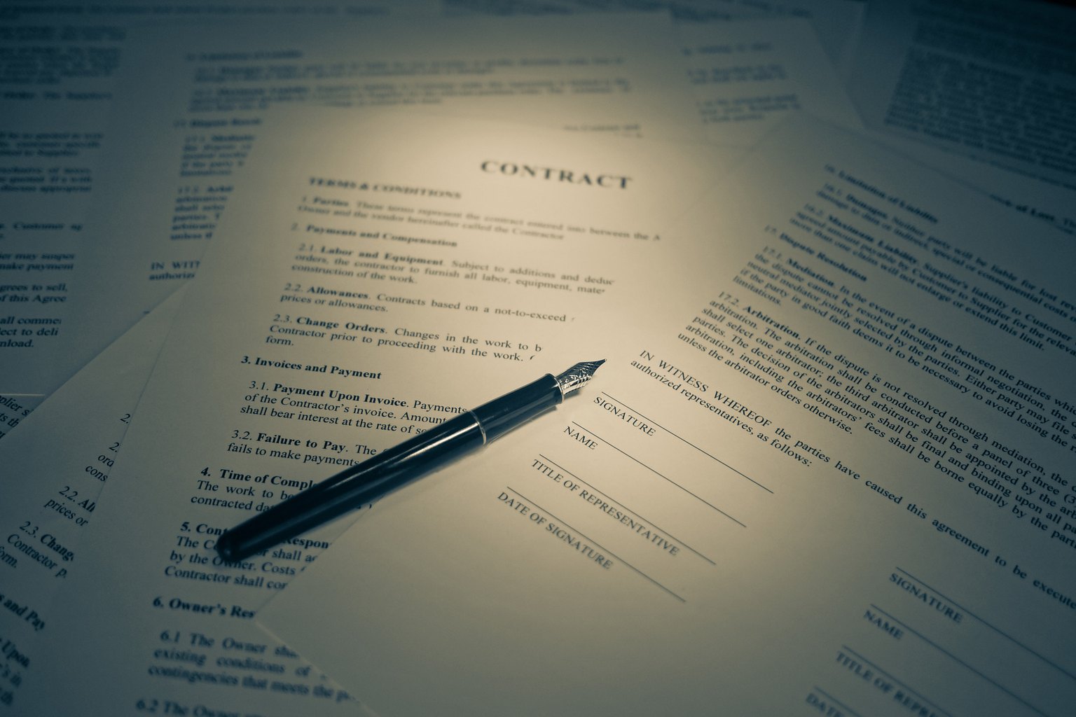 A printed contract page headed CONTRACT resting on a desk under a lamp, with signature, title of representative and date of signature lines, and a fountain pen lying across the page