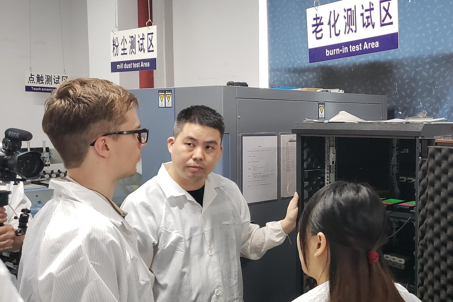 Visitors in white coats standing at a burn-in test cabinet in an electronics factory, with bilingual signs for dust test area and burn-in test area above the equipment
