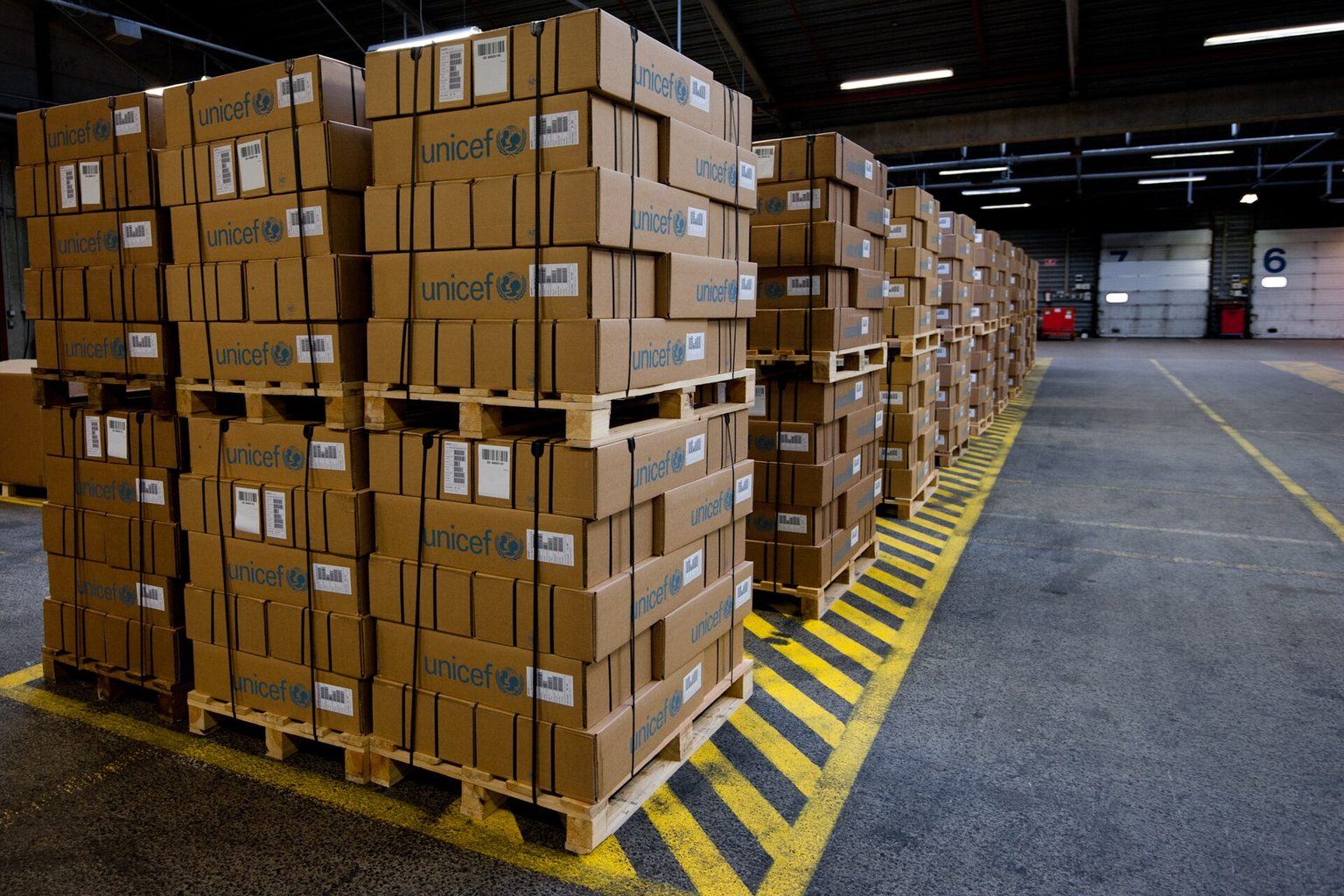 Stacks of sealed brown shipping cartons on wooden pallets with printed labels, lined up inside a distribution warehouse beside a loading door