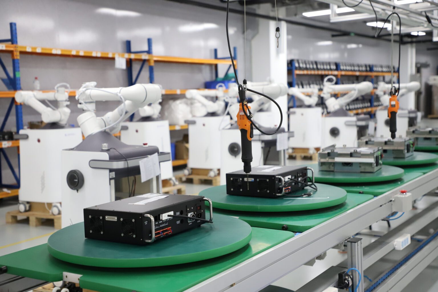 A clean electronics assembly line with white robotic arms working over green rotating turntables, with electronic units on each station and component shelving along the wall behind