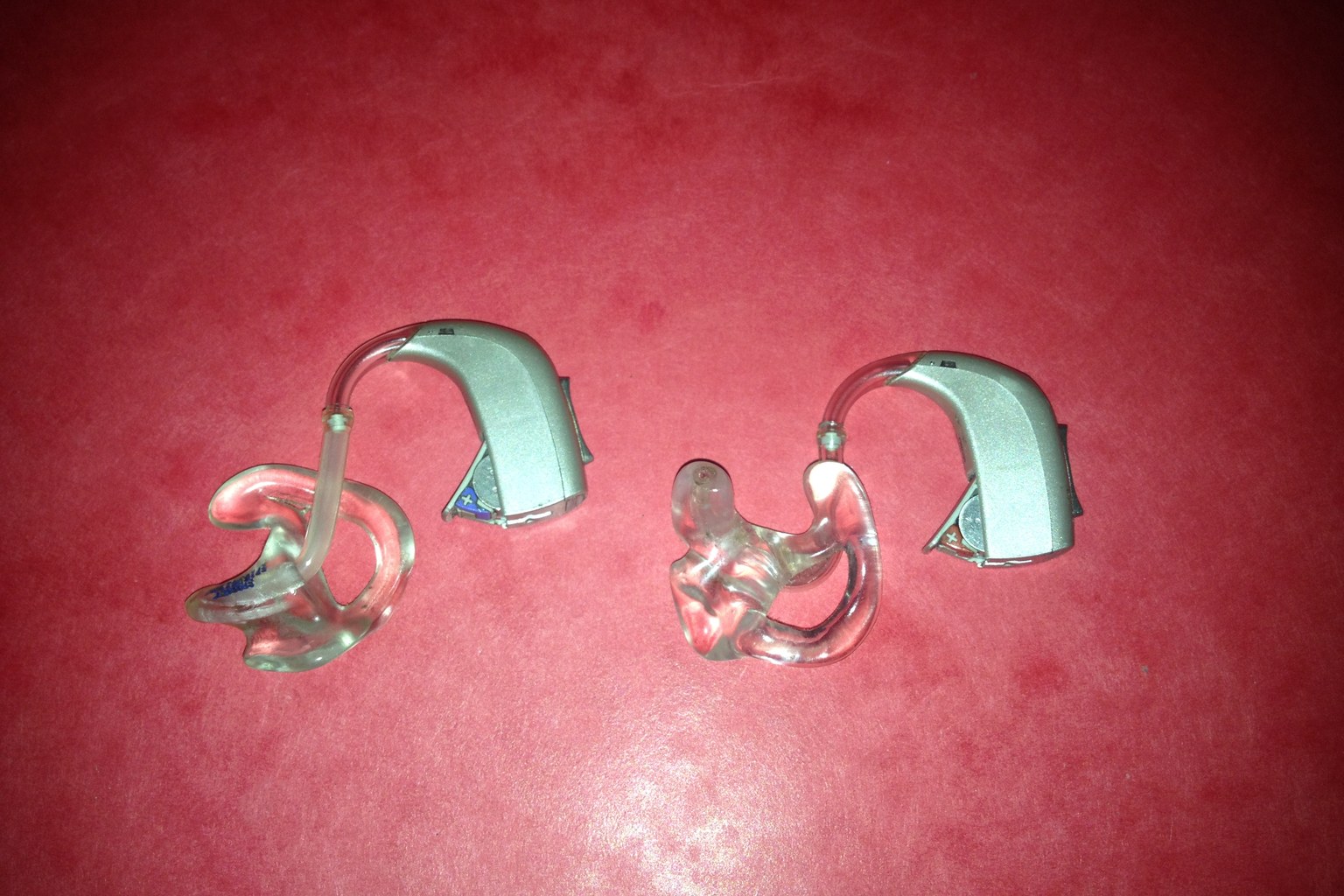 A pair of behind-the-ear hearing aids on a red background, showing the curved housing that hooks over the ear and the clear moulded tube that carries sound into the canal