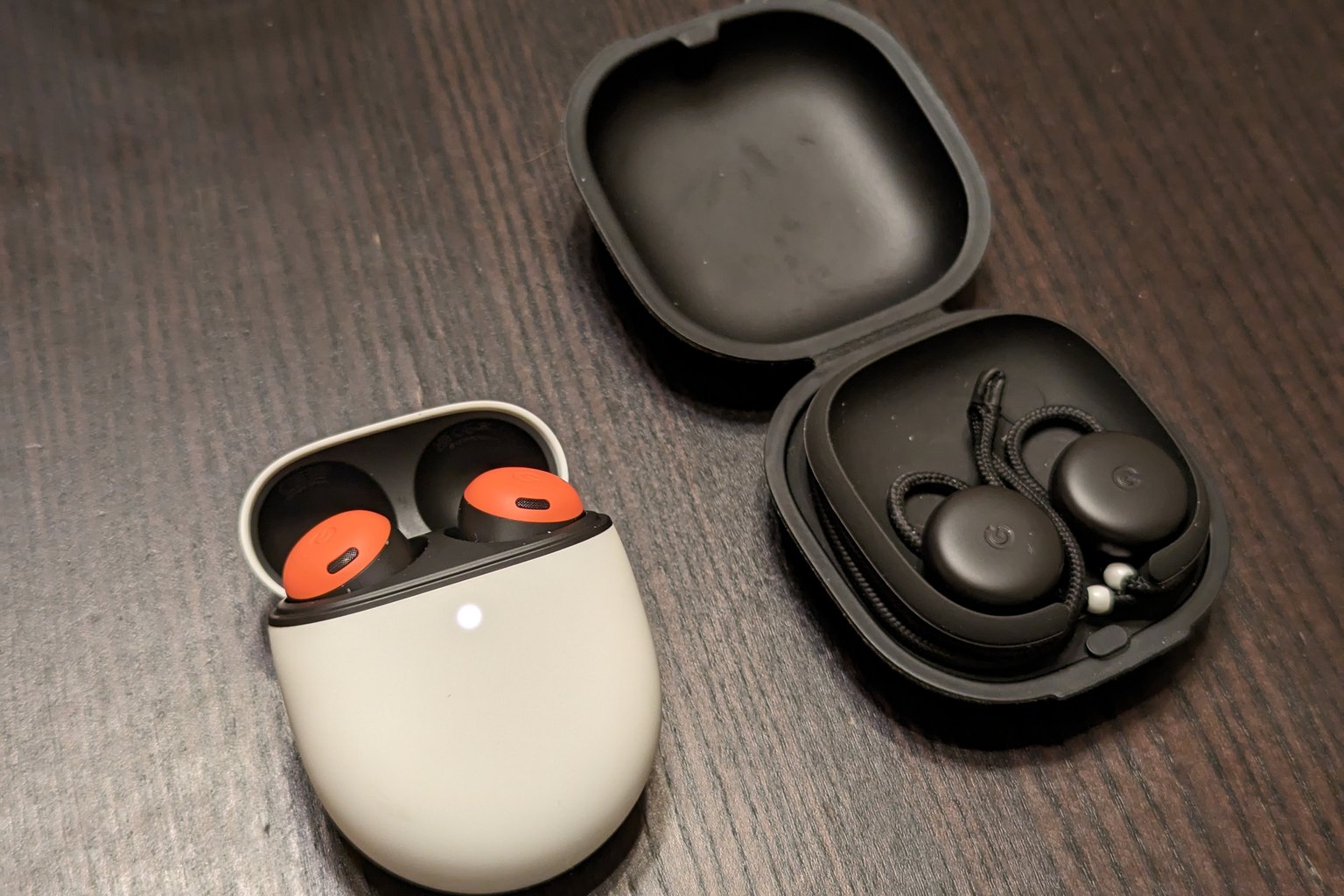 Two open wireless earbud charging cases on a dark wooden table, one holding coral earbuds and one holding a black pair with a connecting cord
