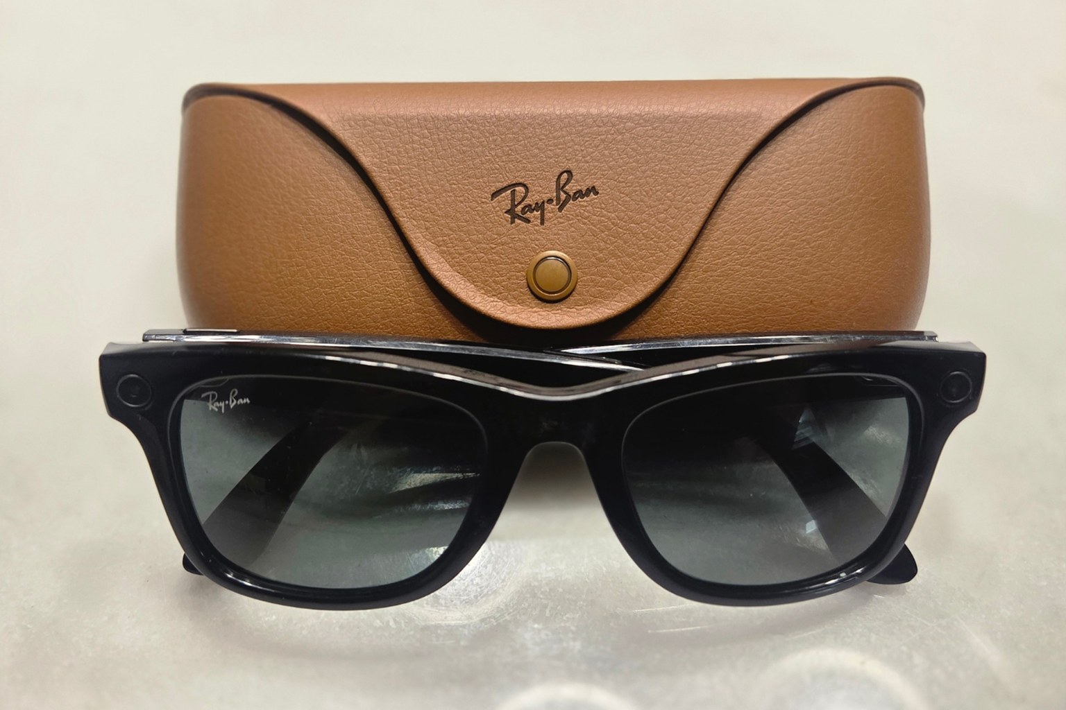 A pair of black Ray-Ban Meta smart glasses resting in front of their tan leather case on a white table
