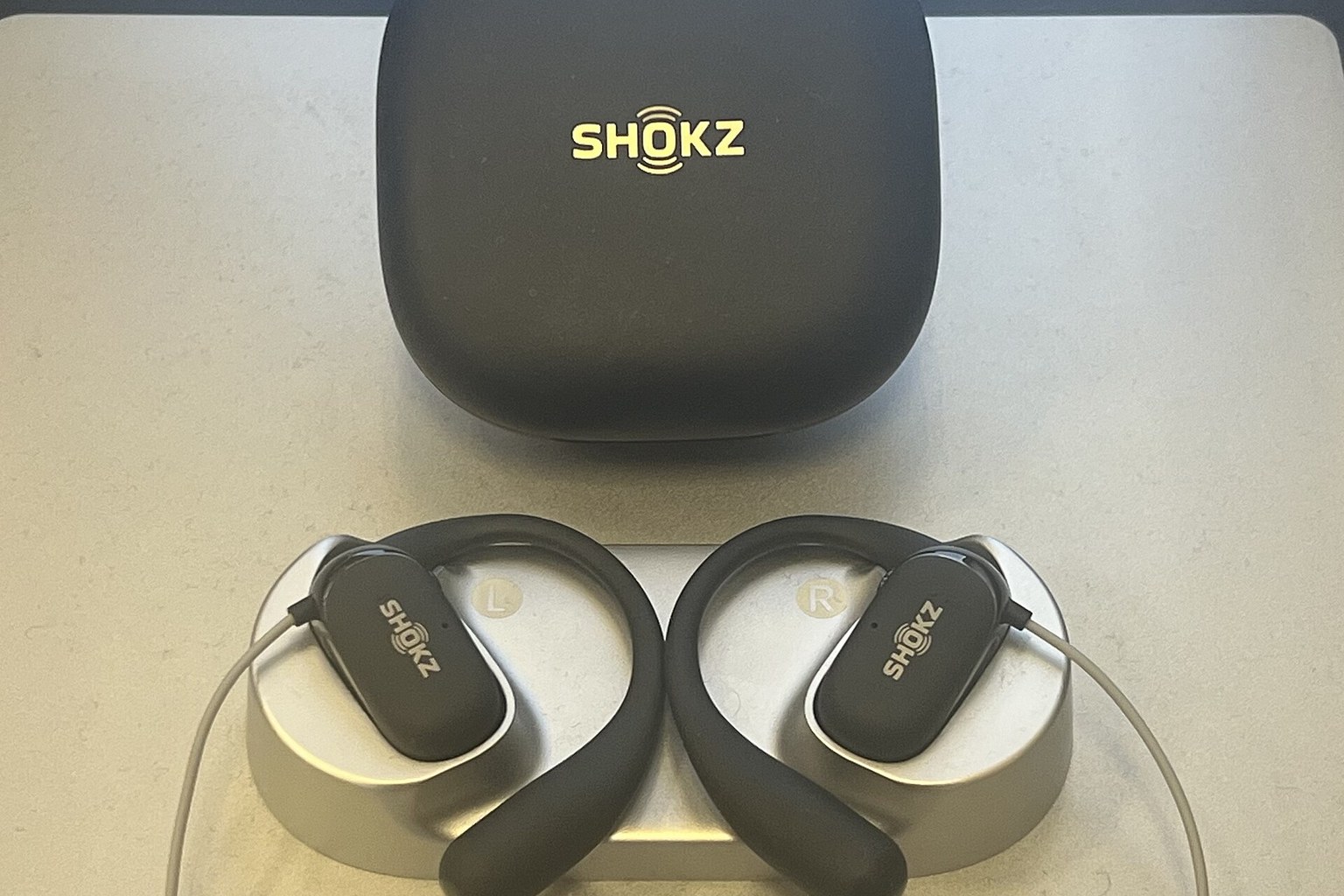 Two hook-style open-ear earbuds with flexible arms resting below their closed charging case