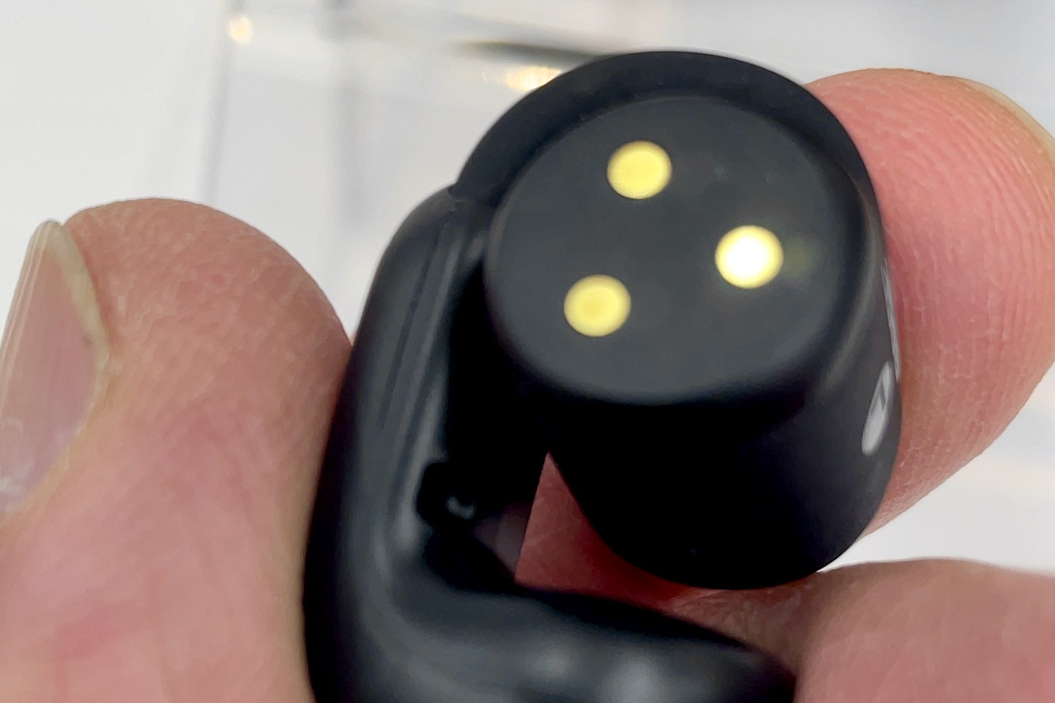 A single black open-ear clip earbud held between fingers, showing three gold charging contacts on the battery barrel