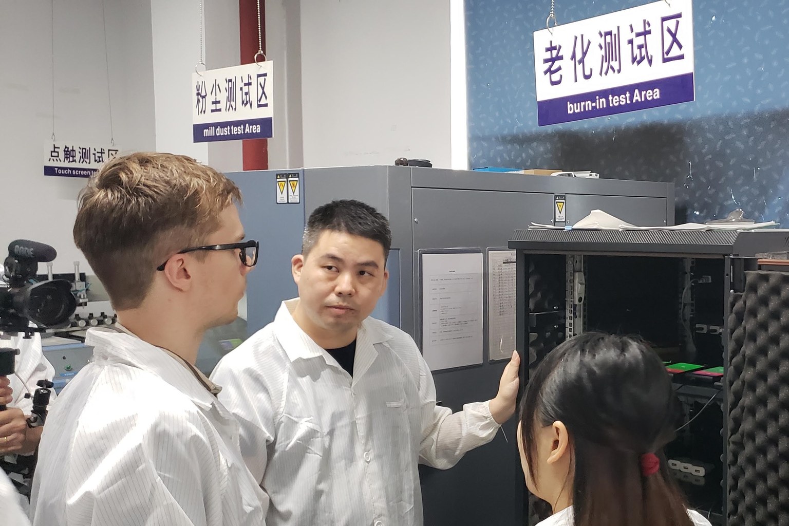 Three people in white coats standing at a burn-in test cabinet in an electronics factory, with bilingual signs for dust test and burn-in test areas
