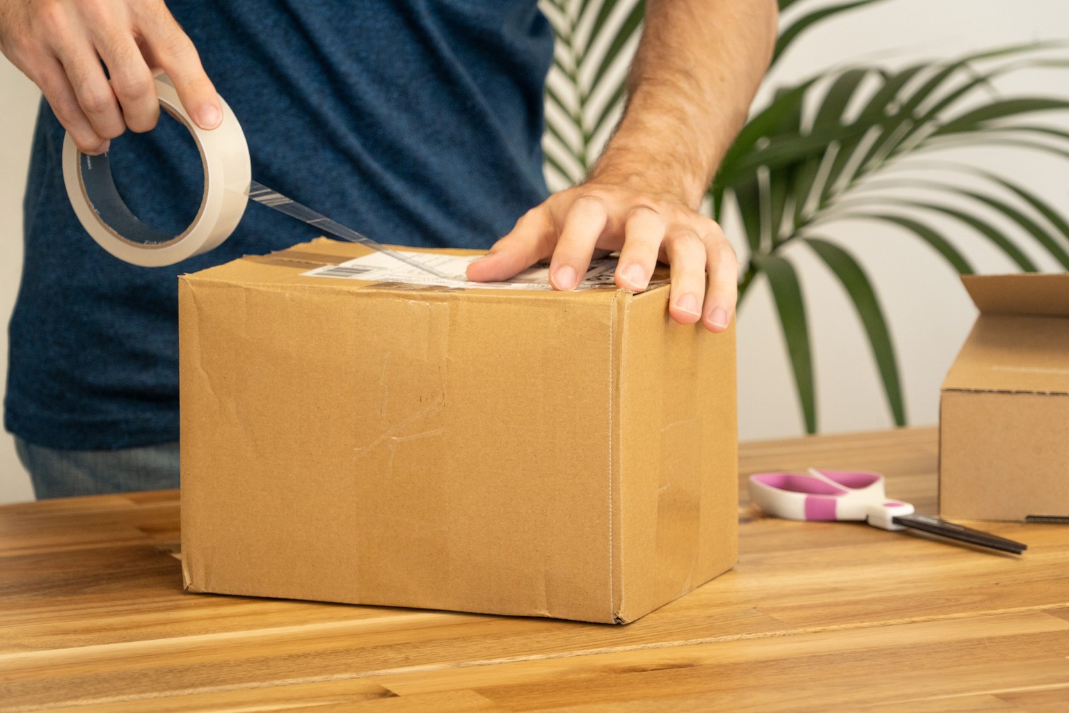 Hands sealing a brown corrugated carton with clear tape and pressing a printed shipping label onto the top face