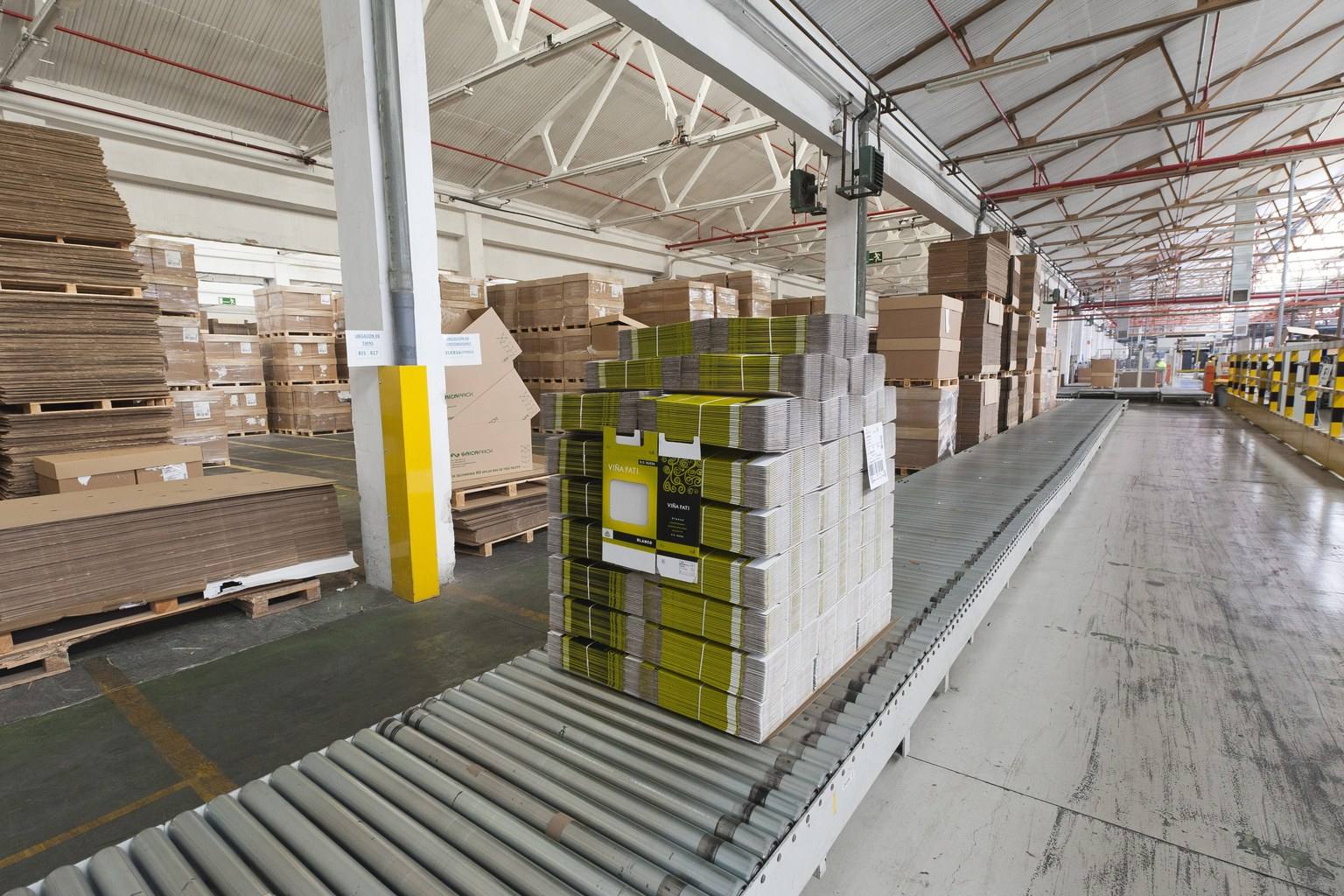 Palletised stacks of printed cartons and corrugated board moving along a roller conveyor inside a packaging plant