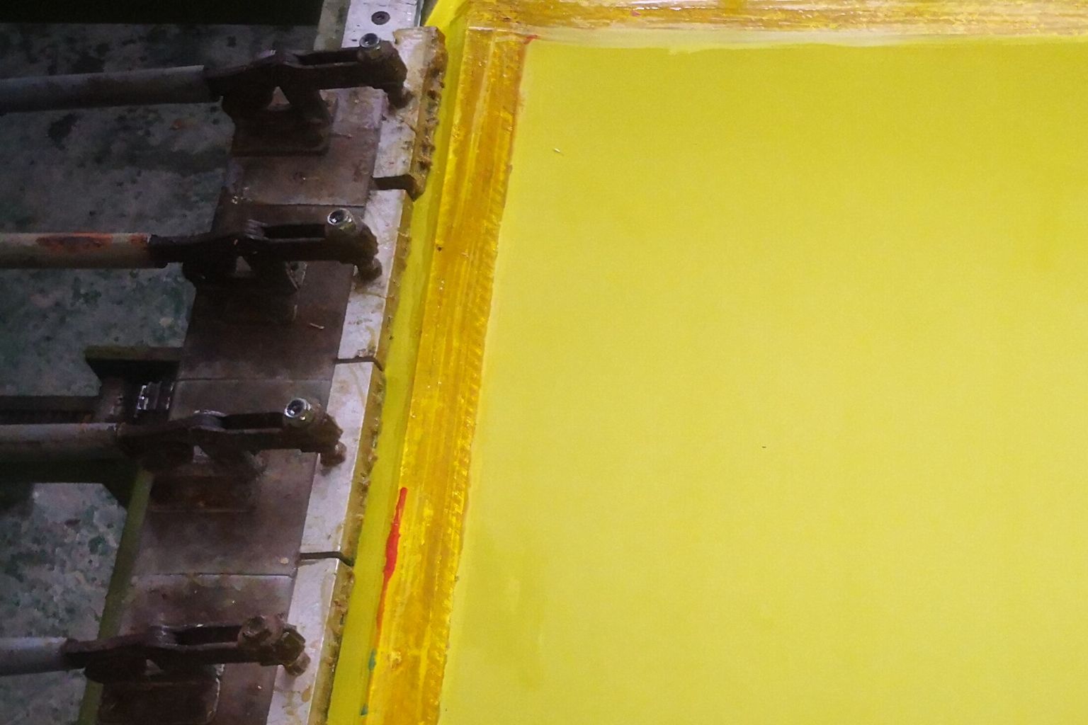A rectangular silk screen printing frame with yellow photosensitive emulsion stretched taut, held in a steel clamp frame on a workshop bench