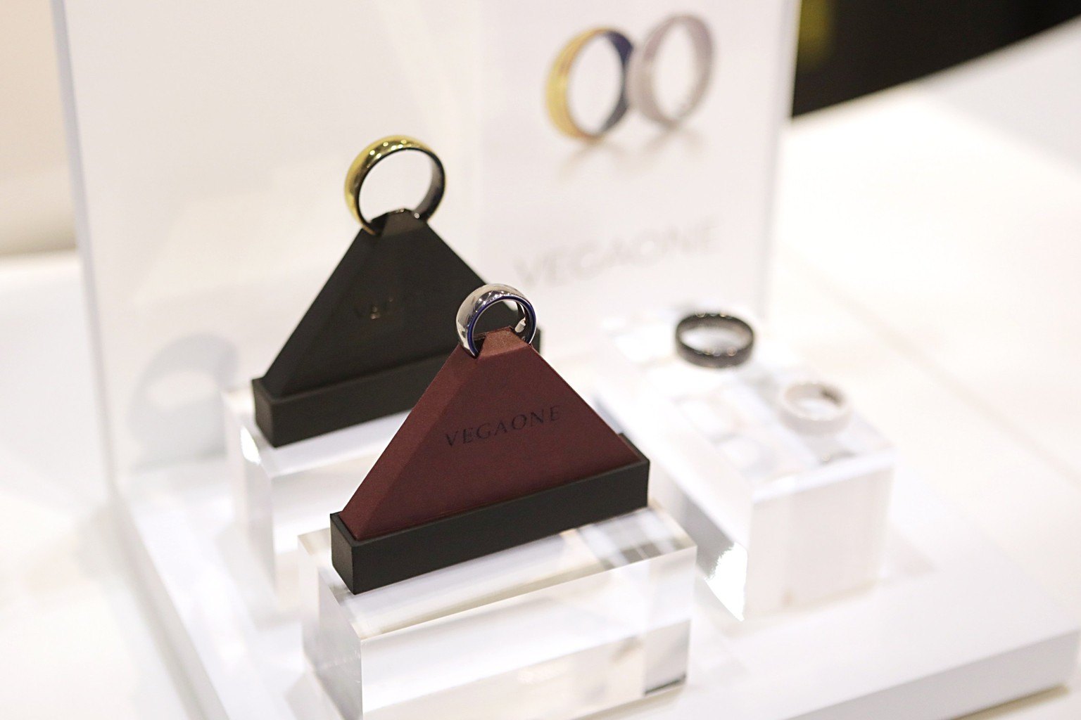 Three smart rings in gold, black and purple finishes displayed on angled stands at a trade show, with a product backdrop behind them
