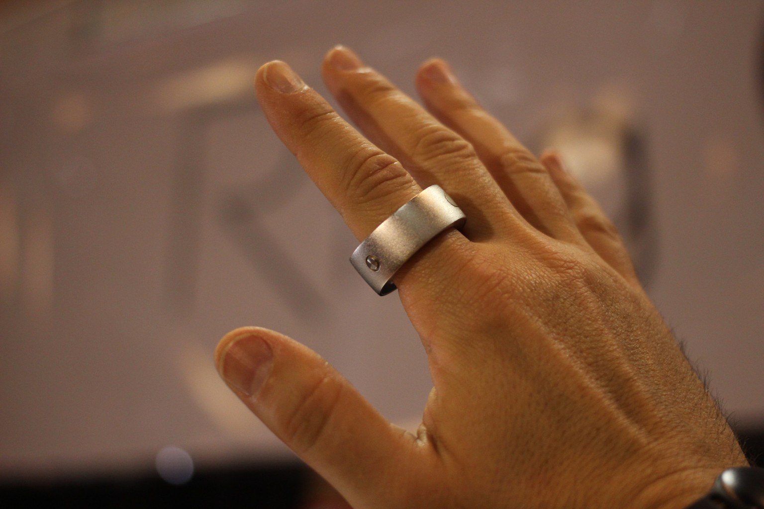 A hand wearing a brushed metal ring-shaped gesture controller, photographed at a hardware fair with a blurred exhibition hall behind it