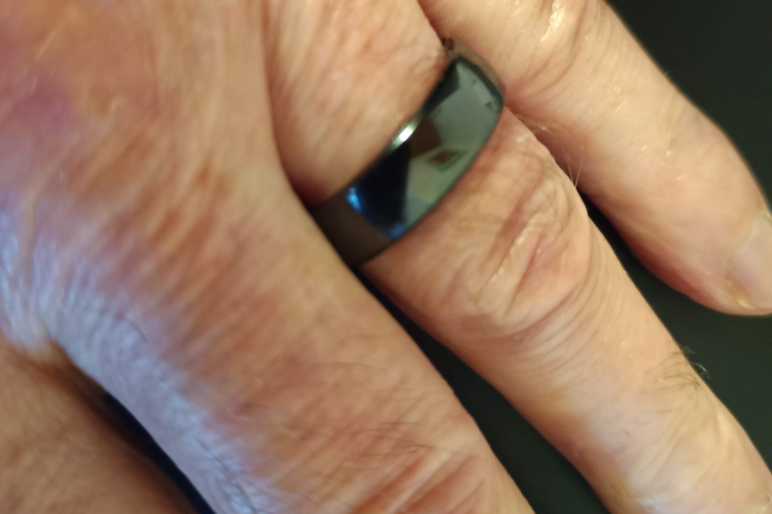 Close-up of a black smart ring worn on a finger, with the sensor band facing the skin and a dark background behind the hand